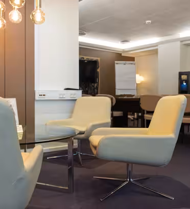 Moderne møterom med lyse beige stoler rundt et glasssalongbord. Varme overlys henger over. I bakgrunnen er det en TV, flippover, og flere sitteplasser ved et langbord. Nøytrale toner skaper en profesjonell atmosfære.