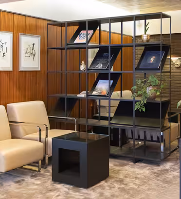 Moderne stue med to beige lenestoler, et svart geometrisk sidebord og dekorative hyller med bøker og planter. Bakgrunnen har trepanelvegger og innrammede kunstverk, som skaper en stilig og komfortabel plass.