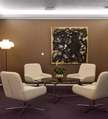 Et moderne møterom med fire beige stoler rundt et sirkulært glassbord. Et stort abstrakt maleri henger på en brun vegg. To stående lamper lyser opp rommet, og en potteplante står på bordet. En flatskjerm-TV er montert på venstre vegg.